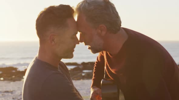 Happy caucasian gay male couple sitting on car playing guitar and kissing at the beach