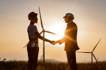 inspector using digital tablet during meeting outdoors and shaking hands. Two partners standing on field with wind turbines during amazing sunset.