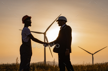 inspector using digital tablet during meeting outdoors and shaking hands. Two partners standing on field with wind turbines during amazing sunset.