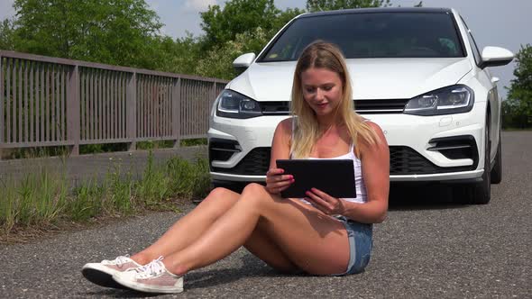 A Young Beautiful Woman Sits on a Road and Works on a Tablet - a White Luxurious Car