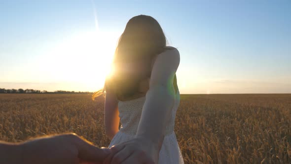 Young Woman Holding Male Hand and Walking on Endless Wheat Field at Bright Sunset Background