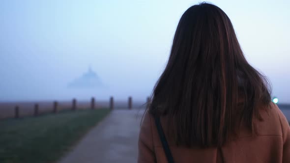 Cinematic Rear View of Beautiful Young Tourist Woman Walking To Sunrise Foggy Mont Saint Michel