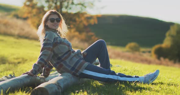 Young attractive blonde woman resting on wooden log in green meadow at sunset.