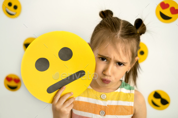 tle girl, sad with a pout, holds a cardboard sad emoji in her hands. Background with emojis of different emotional states: sad, in love, joy, happy