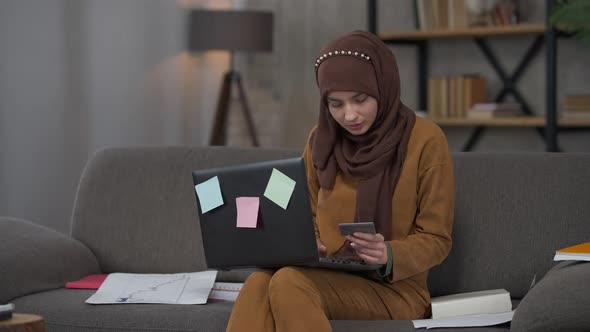 Serious Beautiful Middle Eastern Woman in Hijab Holding Business Card Typing on Laptop Keyboard