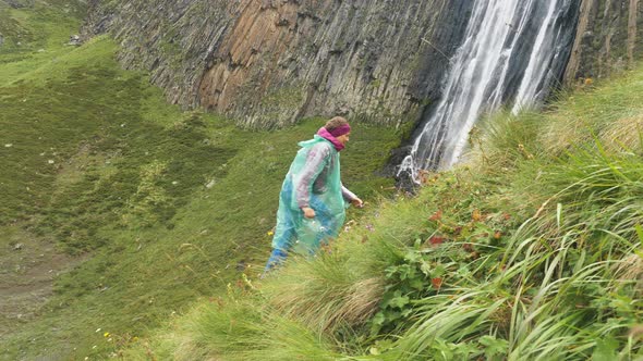 Sports Woman Traveler in a Raincoat Runs Up the Green Mountain Slope Against the Backdrop of a