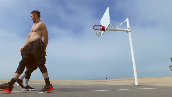 Two men play one-on-one basketball hoops on a beach court.