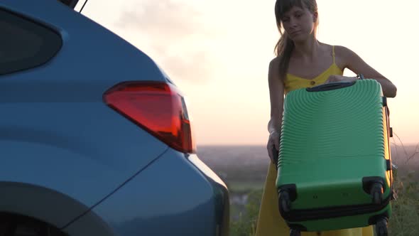 Young woman in yellow dress putting green suitcase in car trunk. Travel and vacations concept.