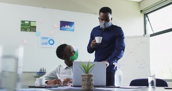 African american man and african american woman wearing face masks discussing over laptop in meeting