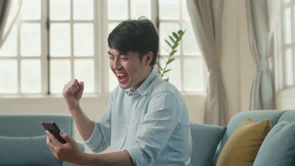 Asian Male Using Smartphone And Celebrating In Living Room