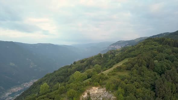 Shooting in flight over the mountains covered by green trees with a city in the valley