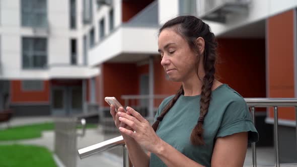 Smiling Woman Is Reading Message in Cell Phone, Standing Outdoors Near High Apartment House