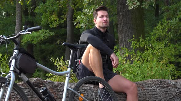 A Young Handsome Cyclist Sits on a Log Next To His Bike in a Forest and Looks Around