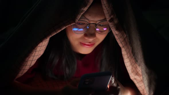 Happy Asian woman using smartphone underneath duvet in the dark on bedroom at home.