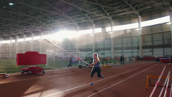 Pole Vaulting Indoors - Young Woman with Pigtails Jumping Over the Bar and Touches the Bar