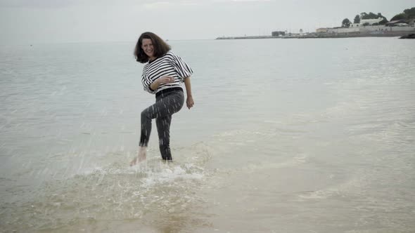 Joyful Beautiful Brunette Woman Kicking Ocean Waves and Smiling