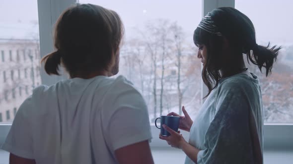 Happy Young Multiracial Couple Drinking Morning Coffee in Front of the Window and Watching Park