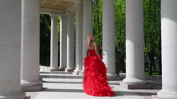 a Blonde with Big Breasts in a Red Fluffy Dress and with a Rose Poses Among the Columns