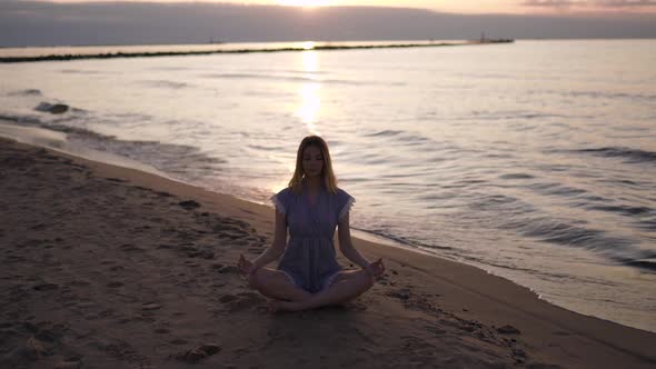 Caucasian Blonde Woman Meditates at Sandy Beach Wearing Blue Summer Dress During Sunrise