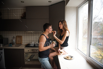 Young millennial couple is sitting on the windowsill near window, drinking latte coffee with cookies