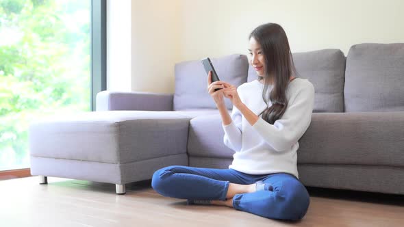 Pretty Asian Woman Playing Online Game While Sitting on the Floor of Living Room on Bright Day, Full