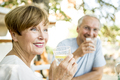 Smiling senior woman drinking glass of water with husband in background - PhotoDune Item for Sale
