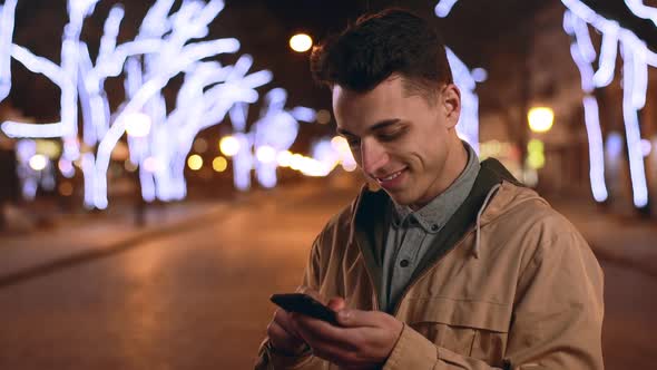 Portrait of European Guy 20s Laughing and Holding Mobile Phone While Using Internet During Night