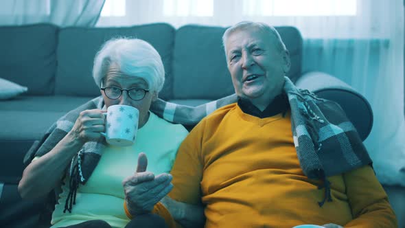 Happy Old Couple Laughing Watching Tv and Drinking Hot Beverage Covered with Blanket