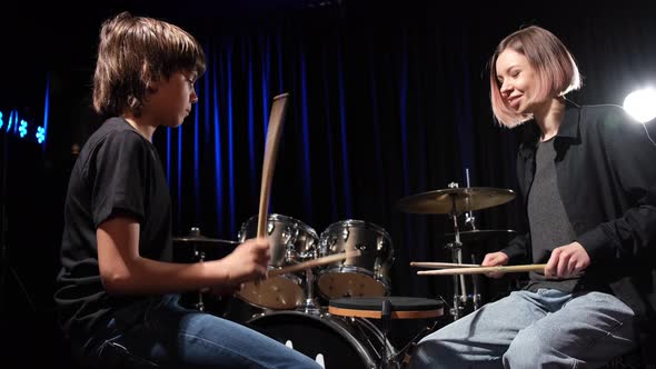 Young Caucasian Woman Teaches a Boy to Play the Drums in the Studio on a Black Background alt