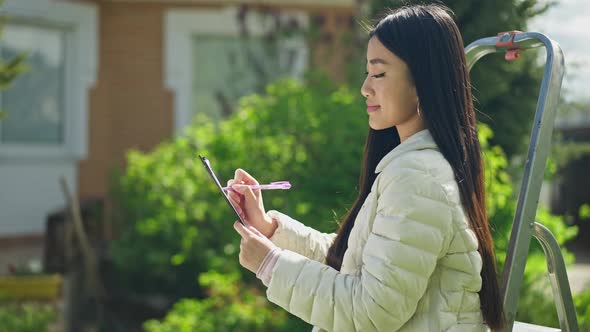 Side View Portrait of Confident Female Asian Gardener Sitting on Ladder Writing in Sketchpad