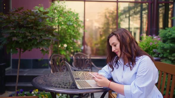 The Woman Sits on an Outdoor Terrace and Works on a Laptop and a Smartphone