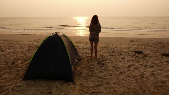 Young woman standing in front of the tent on the beach and enjoying sun over sea in morning