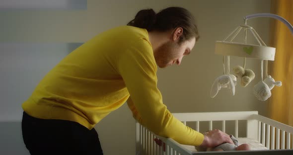 Happy Young Father Strokes Newborn Baby Lying in a Child Crib