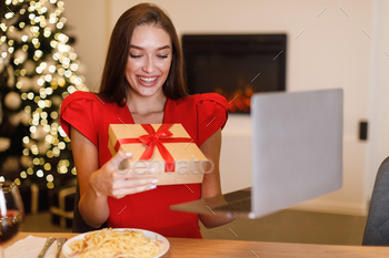 ss celebrating holiday online, holding gift box making video call with boyfriend, husband or family, sitting at table with laptop computer at home