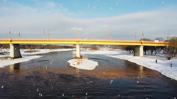 Yellow bridge over river Neris in Kaunas city during snowfall, aerial low angle fly backward view