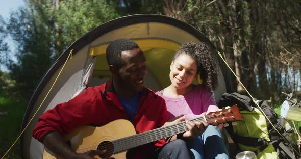 Smiling diverse couple sitting in tent and playing guitar in countryside