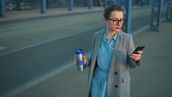 Woman in a Coat Walking Around the City in the Early Morning Drinking Coffee and Using Smartphone