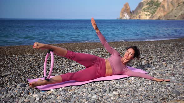 A Young Woman with Black Hair Doing Pilates with the Ring on the Yoga Mat Near the Sea on the Pebble