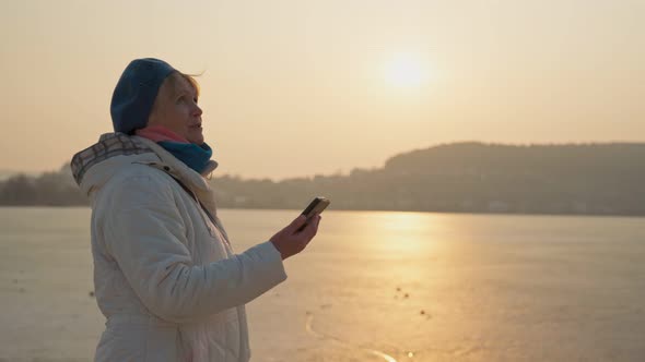 An Older Woman Pretty of Retirement Age on the Shore of the Lake Communicates Via a Smartphone By