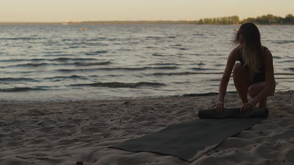 Young Woman Finished Training and Rolls Away a Yoga Mat Against a Beautiful Evening Lake