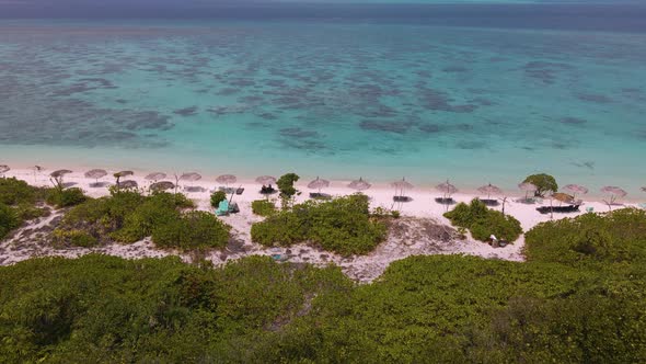 Drone flight over beach with palm umbrellas towards turquoise ocean water over green trees