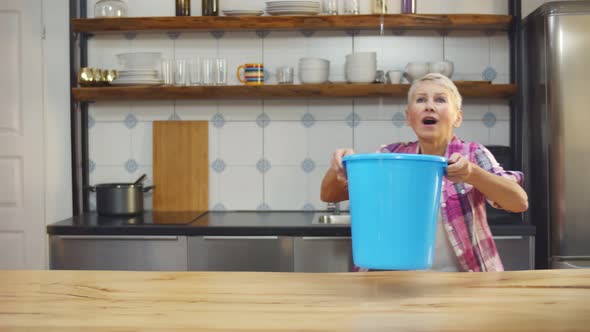 Worried Aged Woman Holding Bucket While Water Droplets Leak From Ceiling in Kitchen