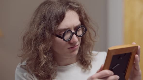 Sad Young Caucasian Man in Retro Eyeglasses with Long Curly Hair Looking at Photograph and Crying