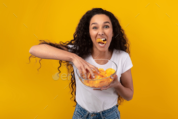 an Enjoying Delicious Potato Crisps Holding Glass Bowl, Posing With Chips In Mouth Isolated Over Yellow Orange Studio Wall. Junk Meal Addiction