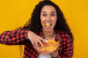 an Enjoying Delicious Potato Crisps Holding Glass Bowl, Posing With Chips In Mouth Isolated Over Yellow Orange Studio Wall. Junk Meal Addiction