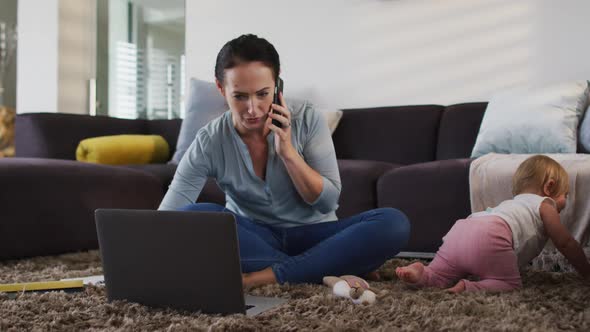 Caucasian mother using laptop and talking on smartphone while baby crawling on the floor at home