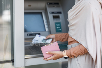tions, payment of financial bills. Unrecognizable young arab female in hijab holds dollars and pink wallet, dials code on atm machine, outdoor