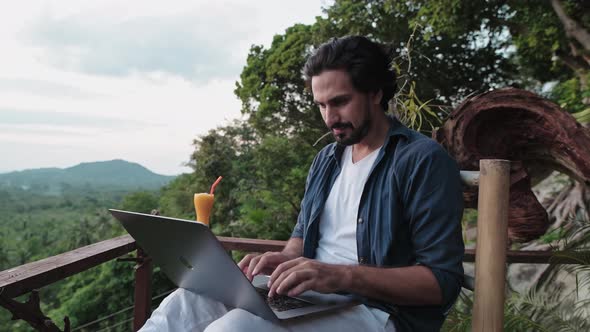 A young, handsome man with a beard works on a laptop in a cafe overlooking an incredible sunset.