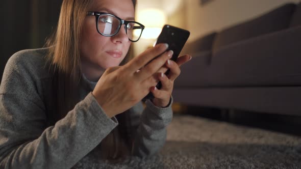 Woman with Glasses Is Lying on the Floor and Using Smartphone in the Evening