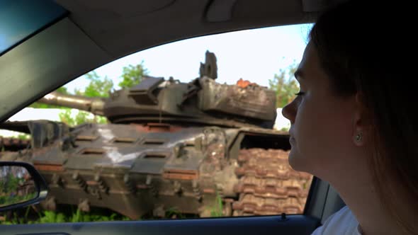 Side View Young Beautiful Woman Sitting in Car Looking at Burnt Tanks in Ukraine Sighing Looking